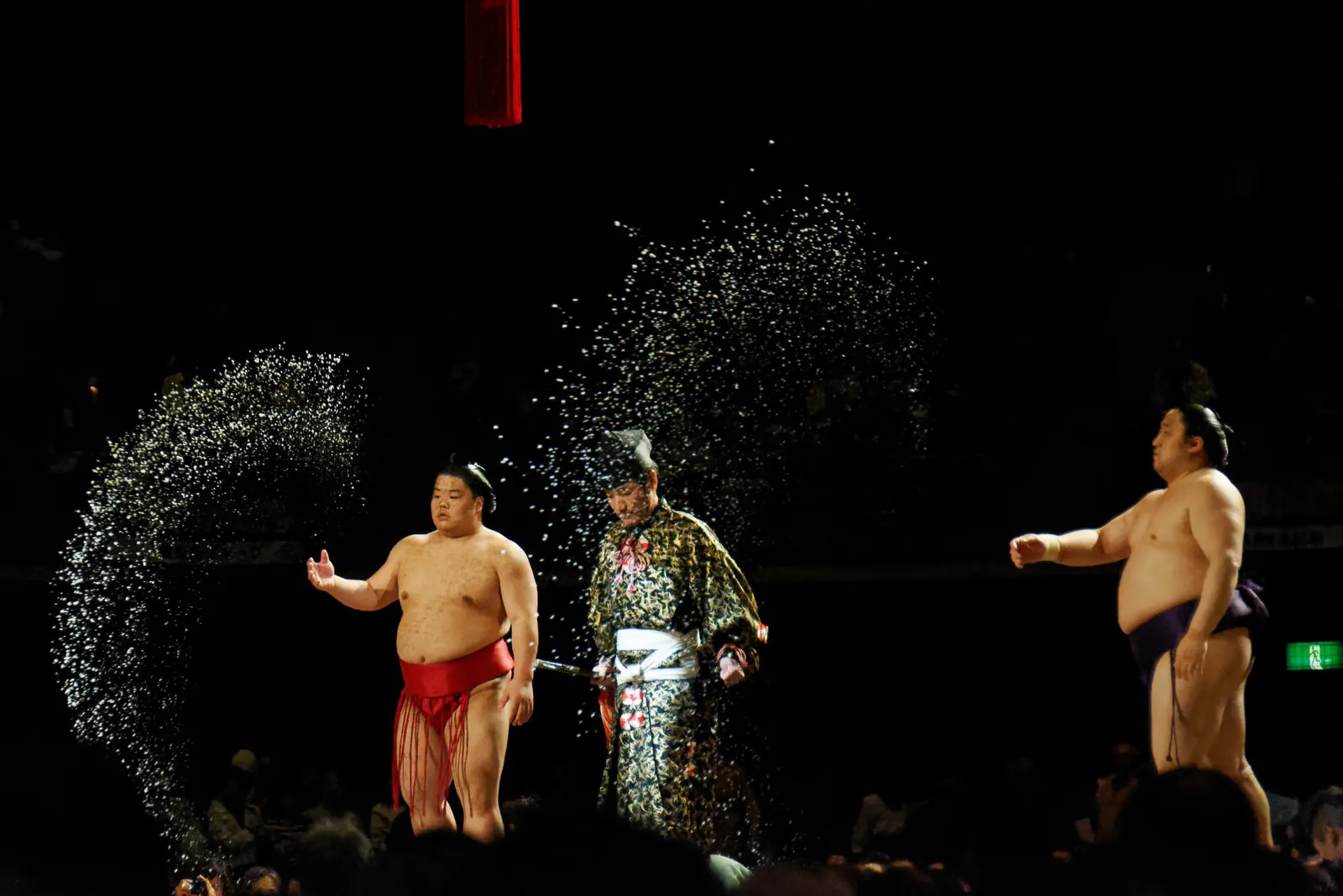 Sumo wrestlers in a dark arena with a spray of white salt hanging in the air around a referee in ceremonial dress