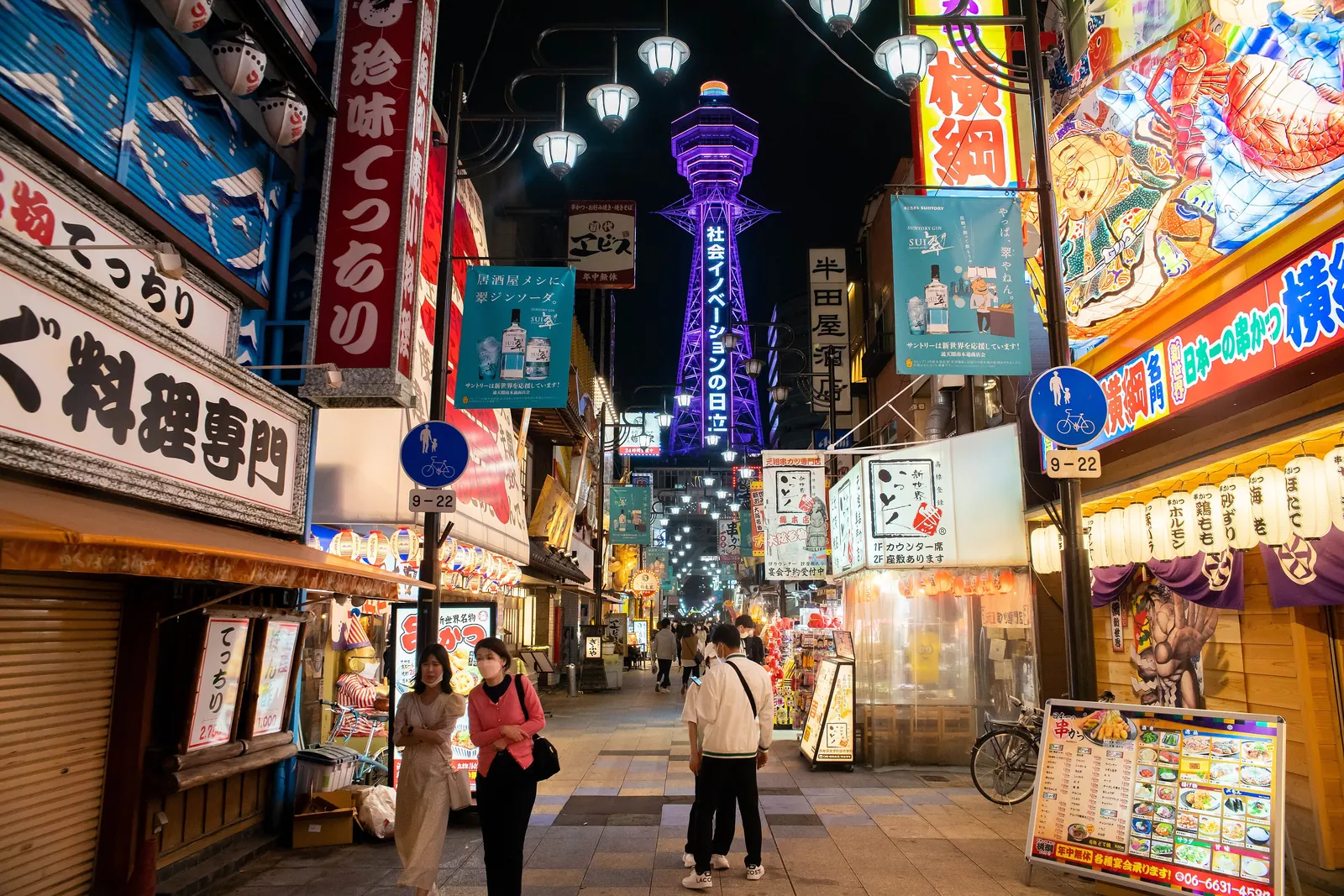 A lit tower glowing purple above a street of neon signs and lanterns in Shinsekai at night