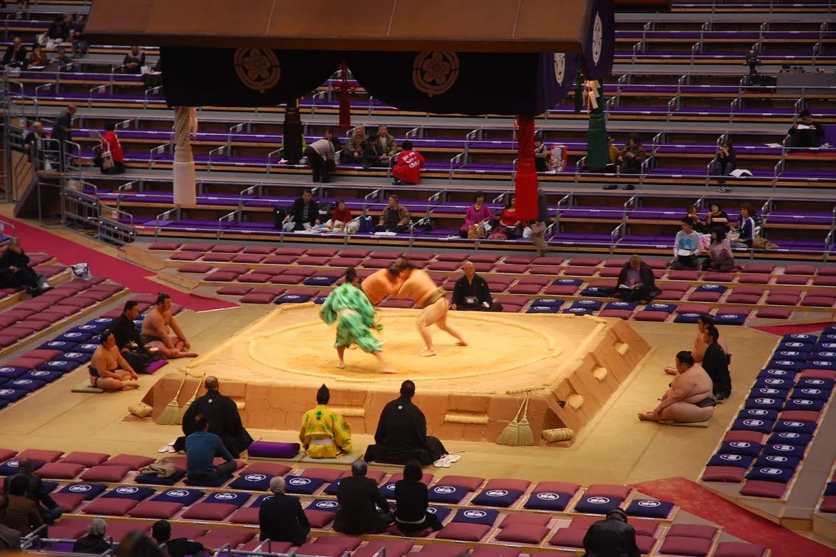A sumo ring on a raised clay platform seen from the seats of an arena, with wrestlers and a referee around it