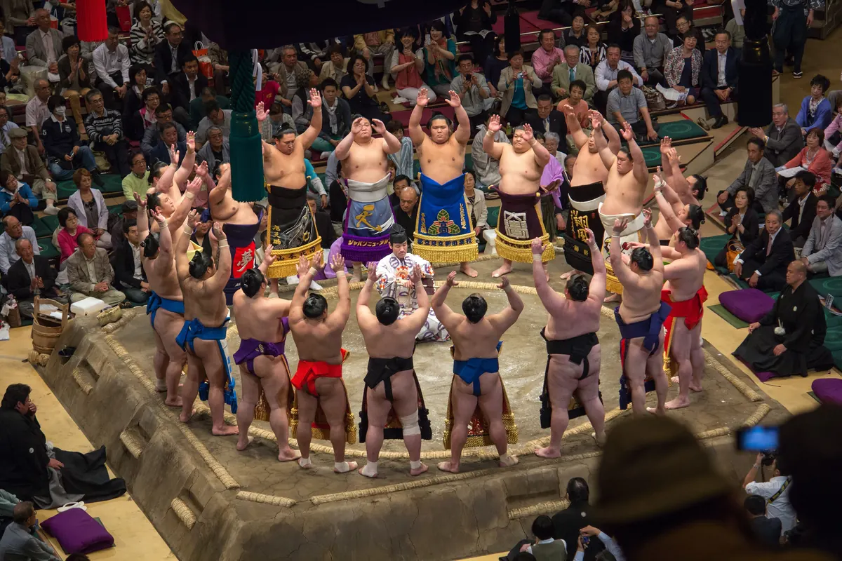 A ring of sumo wrestlers in colourful ceremonial aprons standing on a clay platform with arms raised above their heads