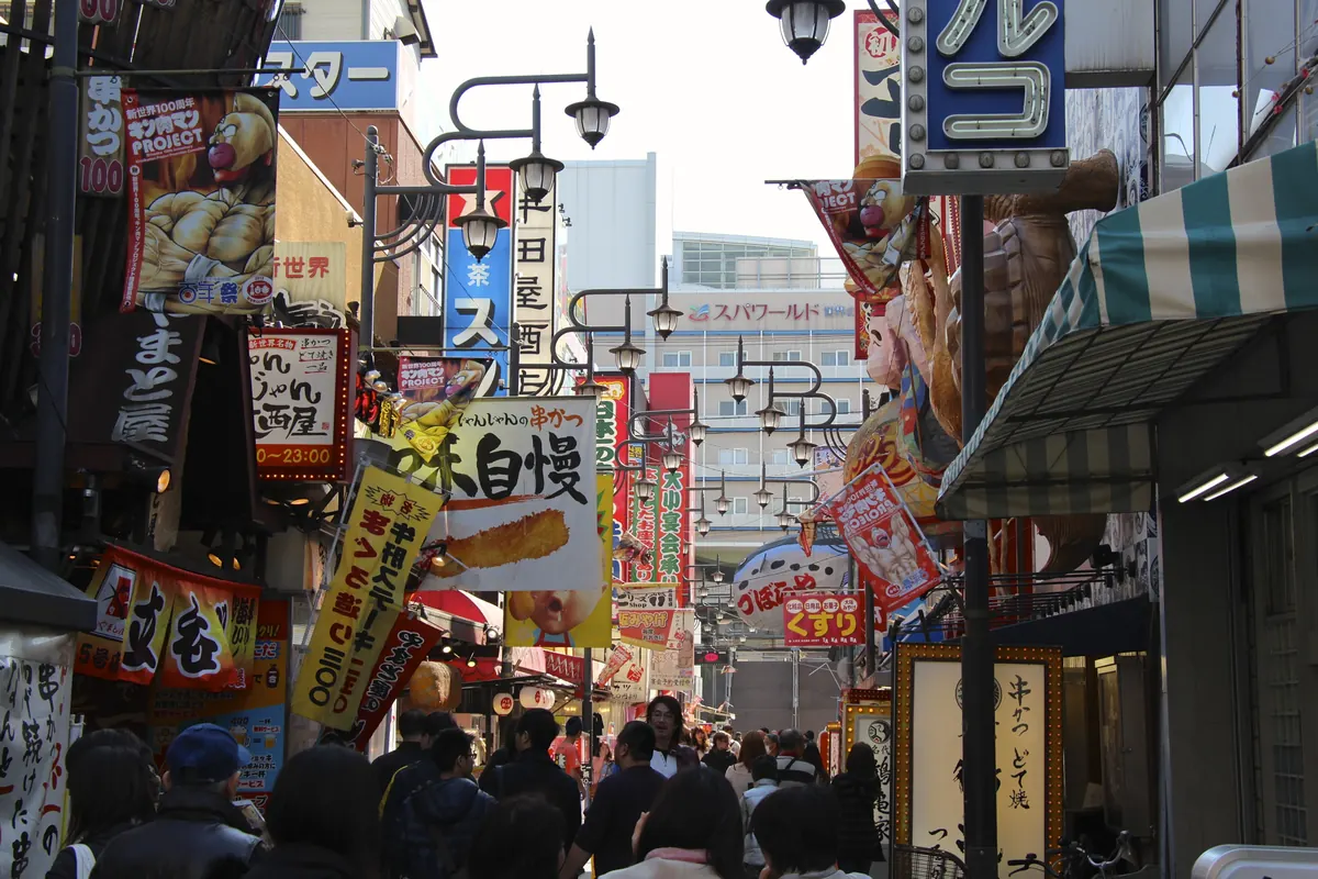 A busy narrow street in Osaka crowded with shop signs, lanterns and food stall banners above the heads of pedestrians